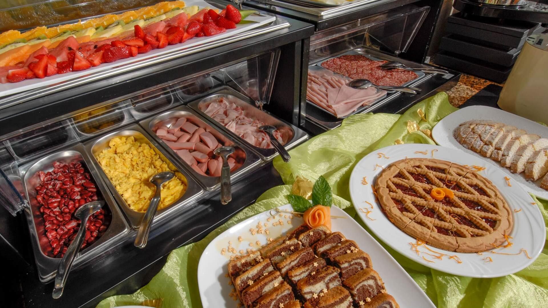 Buffet con frutta, salumi, dolci e uova strapazzate su tovaglia verde.