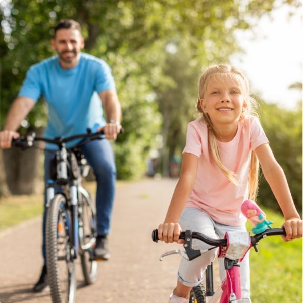Bambina felice in bici seguita da un adulto su una pista ciclabile alberata.