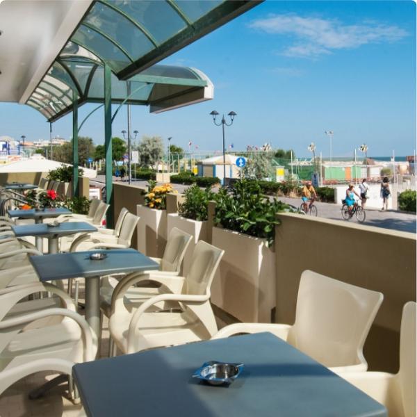 Terrazza con tavoli e sedie, vista mare, sotto una tettoia trasparente.
