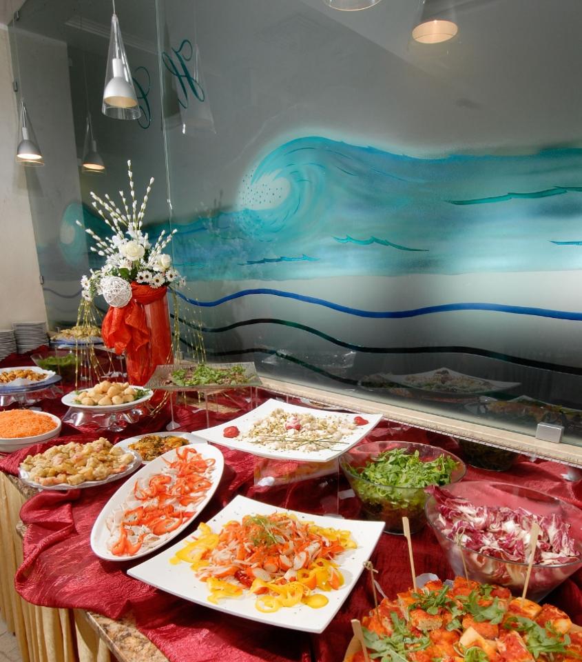 Buffet elegante con piatti vari, decorazioni floreali e tovaglia rossa su sfondo marino.
